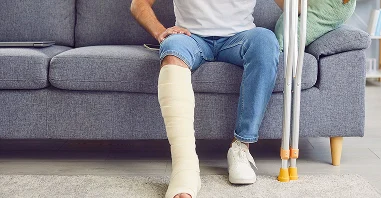A person sits on a couch with a heavily bandaged leg and crutches nearby, wearing casual clothes and looking relaxed.